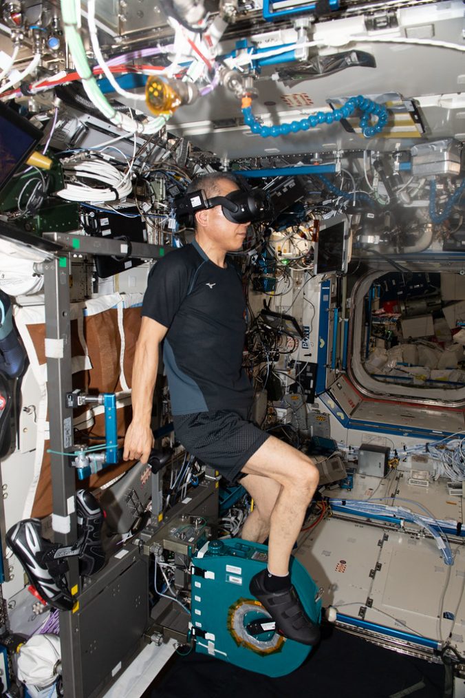 Astronaut Satoshi Furukawa pedals on an exercise cycle while wearing VR goggles